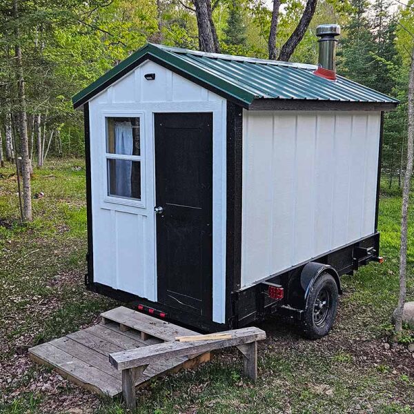 White and black mobile wood-fired sauna on a trailer parked in a wooded Minnesota clearing.