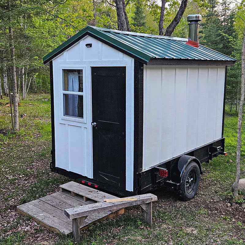 Mobile Sauna with Wood Fired Stove – Delivered to You White and black mobile wood-fired sauna on a trailer parked in a wooded Minnesota clearing.